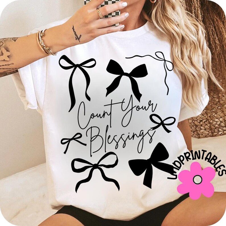 K&ouml;nnte beinhalten: Wei&szlig;es T-Shirt mit schwarzen Schleifen-Illustrationen und dem handgeschriebenen Schriftzug "Count Your Blessings". Das Design umfasst verschiedene Schleifenformen, die &uuml;ber das Shirt verteilt sind. Eine rosa Blume mit dem Text "LNDPRINTABLES" befindet sich unten rechts.