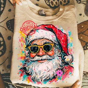 May include: A beige sweatshirt with a vibrant Santa Claus graphic. Santa is depicted wearing yellow sunglasses and a red hat. The design includes the text "THE ORIGINAL" in a circular stamp. The illustration is surrounded by colourful paint splatters.