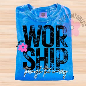Pu&ograve; includere: Una maglietta blu chiaro piegata con la parola "WORSHIP" in grandi lettere nere consumate. Sotto, la frase "through the worry" &egrave; scritta in corsivo. La maglietta ha un'etichetta Comfort Colors.