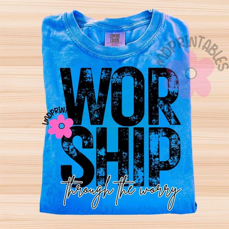 Pu&ograve; includere: Una maglietta blu chiaro piegata con la parola "WORSHIP" in grandi lettere nere consumate. Sotto, la frase "through the worry" &egrave; scritta in corsivo. La maglietta ha un'etichetta Comfort Colors.