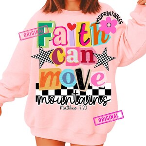 May include: A light pink sweatshirt with the words "Faith can move mountains" in colorful, layered text. The design includes a heart, stars, and a flower. The text "Matthew 17:20" is also included.