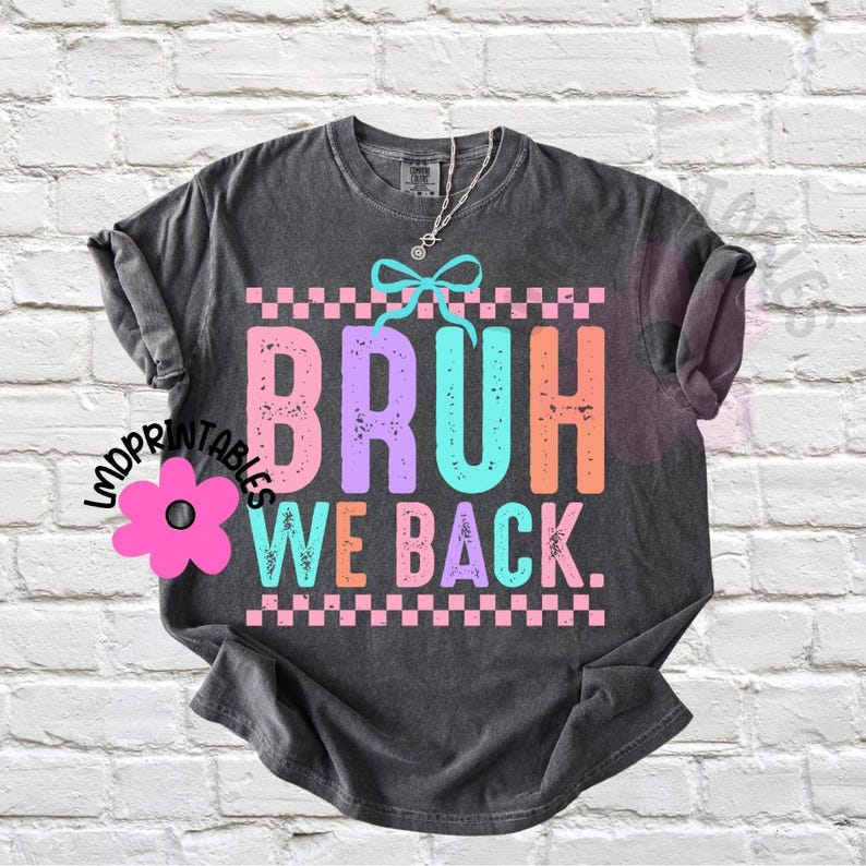 Puede incluir: Camiseta gris oscuro con la frase "BRUH WE BACK" en letras coloridas y desgastadas. El dise&ntilde;o incluye un patr&oacute;n de cuadros y un lazo. Una prenda de vestir informal.