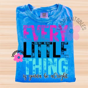May include: A blue Comfort Colors t-shirt with the words "EVERY LITTLE THING is gonna be alright" in pink, black, and blue. The shirt is folded and displayed on a light wood surface.