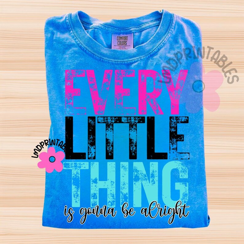 May include: A blue Comfort Colors t-shirt with the words "EVERY LITTLE THING is gonna be alright" in pink, black, and blue. The shirt is folded and displayed on a light wood surface.