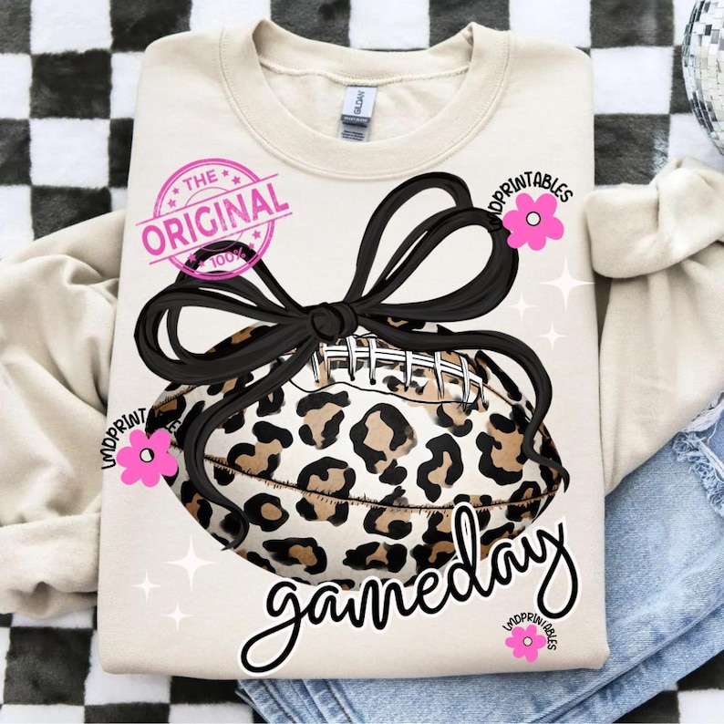May include: Cream-colored sweatshirt with a leopard print football design tied with a black bow. The text "gameday" is written in cursive, with pink flower accents and the text "The Original 100%" in a pink circle.