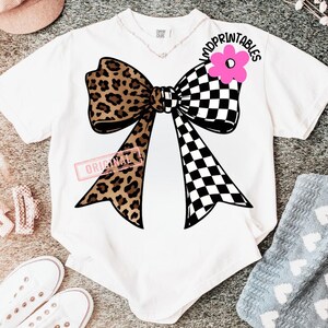 May include: White t-shirt featuring a large bow design. The bow has a leopard print on one side and a black and white checkered pattern on the other. A pink flower and the text "MDPRINTABLES" are above the bow.