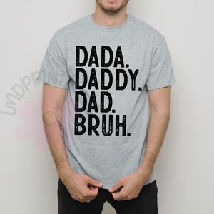 May include: A light gray t-shirt with the words "DADA. DADDY. DAD. BRUH." printed in black, stacked vertically. The shirt is short-sleeved and appears to be made of a soft material.