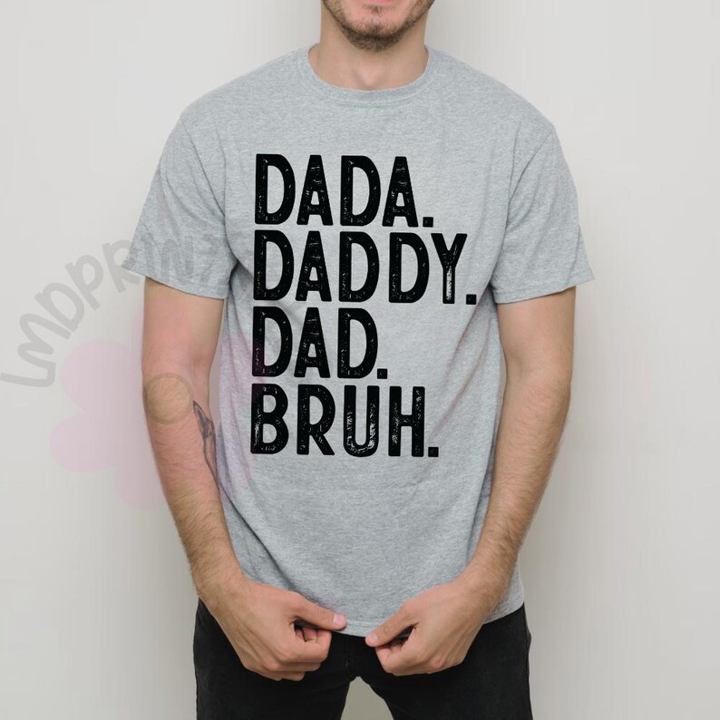 May include: A light gray t-shirt with the words "DADA. DADDY. DAD. BRUH." printed in black, stacked vertically. The shirt is short-sleeved and appears to be made of a soft material.