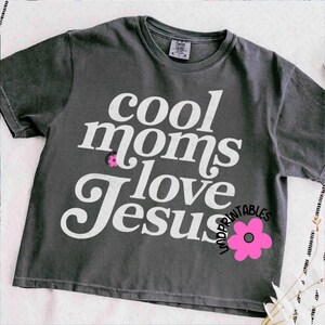 Puede incluir: Camiseta gris oscuro con el texto blanco "cool moms love Jesus" y un gr&aacute;fico de flor rosa. La camiseta es de cuello redondo y parece estar hecha de un material suave. Es una prenda de vestir informal.