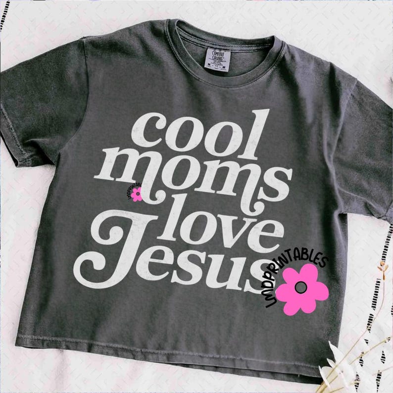Puede incluir: Camiseta gris oscuro con el texto blanco "cool moms love Jesus" y un gr&aacute;fico de flor rosa. La camiseta es de cuello redondo y parece estar hecha de un material suave. Es una prenda de vestir informal.