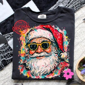 May include: Dark grey t-shirt with a colourful Santa Claus design. Santa wears yellow sunglasses and a red hat. The design includes the text "THE ORIGINAL" and "IMPRINTABLES".