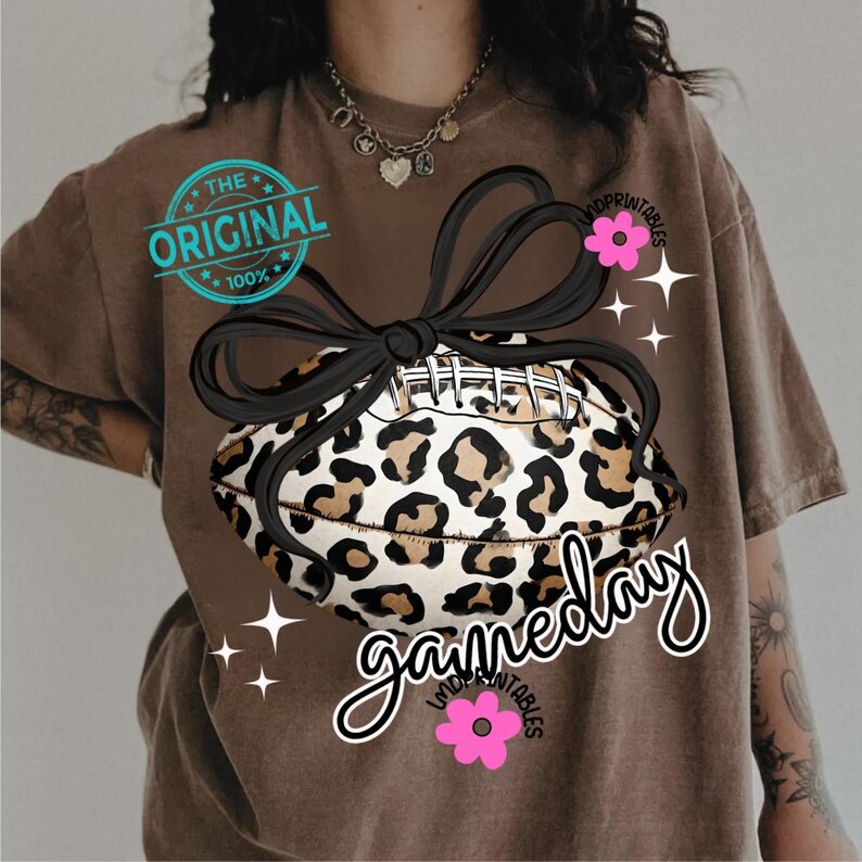 May include: Brown t-shirt featuring a leopard print football with a black bow. The shirt has the text "gameday" in a script font, along with the text "THE ORIGINAL 100%" in a circular stamp.
