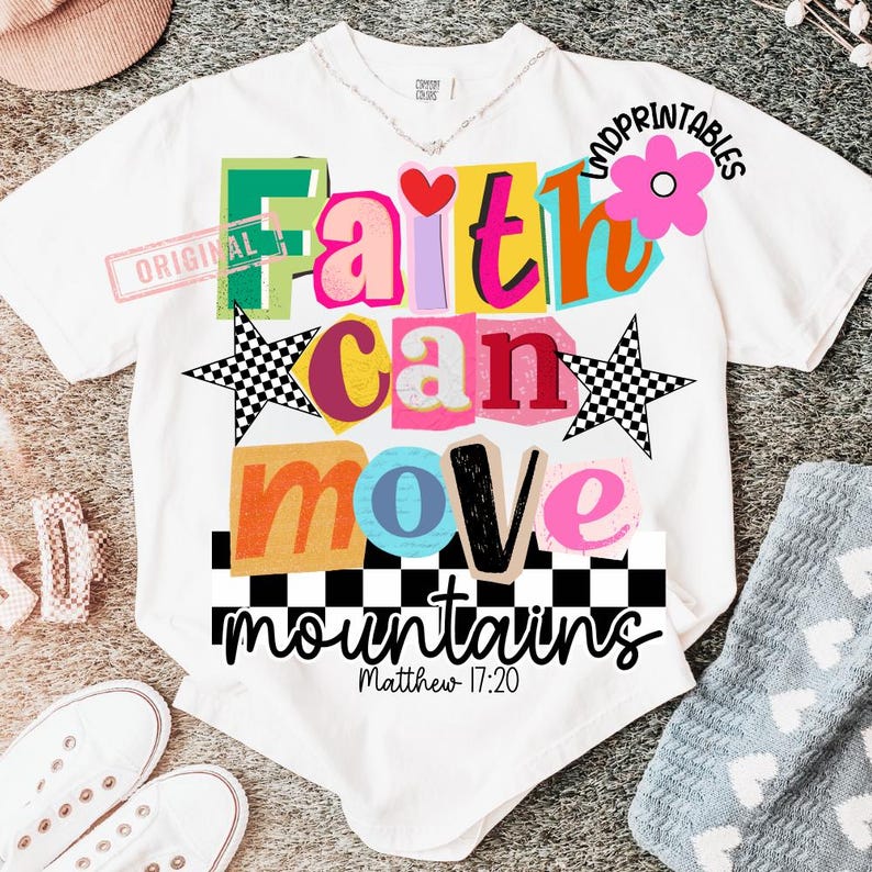 May include: White t-shirt with a colorful graphic design. The design features the words "Faith can move mountains" in various bright colors and fonts, along with two checkered stars and a pink flower. The text "Matthew 17:20" is also included.