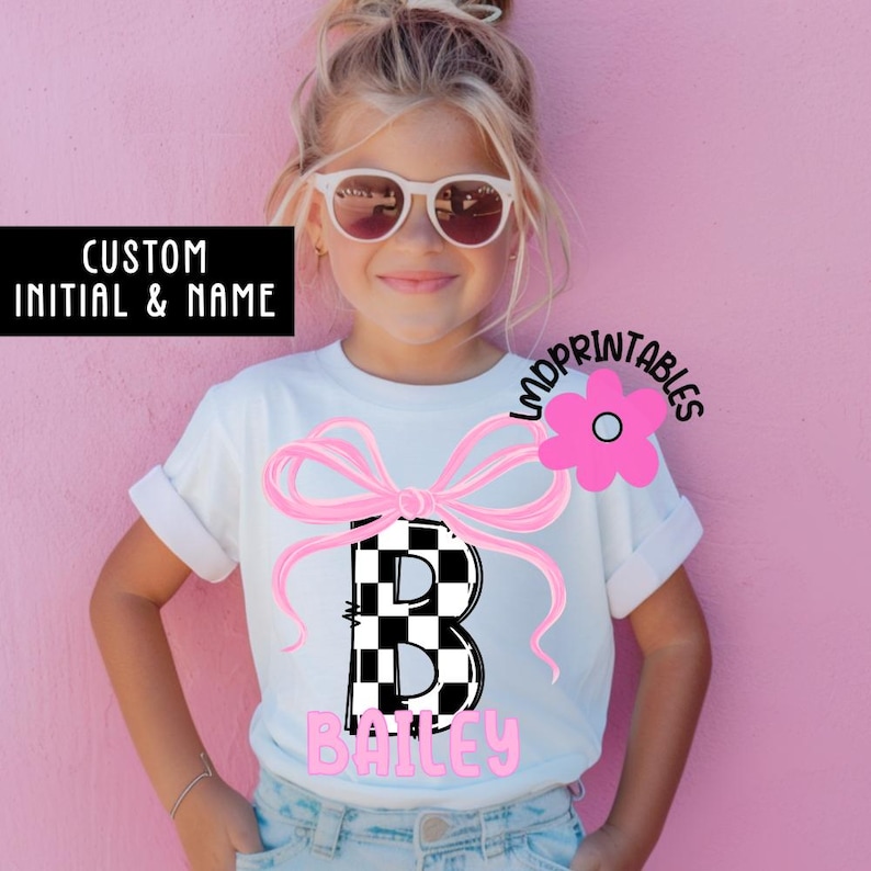 May include: A white t-shirt featuring a large pink bow and a black and white checkered letter "B". The name "Bailey" is printed in pink below the letter. The shirt also has the text "CUSTOM INITIAL & NAME".