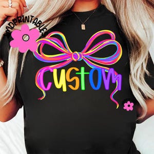 May include: Black t-shirt with a colorful bow design and the word "CUSTOM" in rainbow colors. The bow is made of multiple lines of bright colors. The word "CUSTOM" is in a playful font.