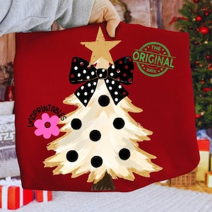 May include: Red Christmas-themed pillow with a cream-colored Christmas tree design. The tree features black polka dots and a black and white polka dot bow. A gold star tops the tree, and the text "THE ORIGINAL 100%" is printed on the pillow.