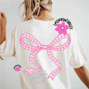 May include: Cream-colored t-shirt featuring a large pink gingham bow design on the back. A pink flower with the text "LMDPRINTABLES" is on the upper right shoulder. The text "THE ORIGINAL 100%" is at the bottom of the bow.