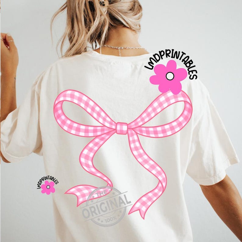 May include: Cream-colored t-shirt featuring a large pink gingham bow design on the back. A pink flower with the text "LMDPRINTABLES" is on the upper right shoulder. The text "THE ORIGINAL 100%" is at the bottom of the bow.