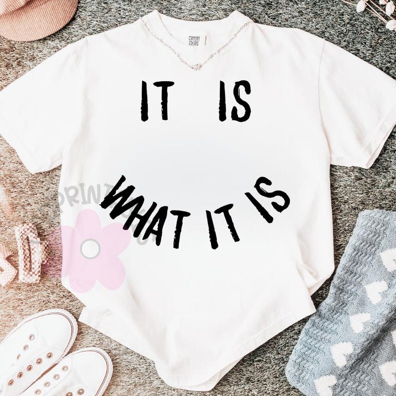 May include: White t-shirt with the text "IT IS WHAT IT IS" in black. The shirt has a crew neck and short sleeves. The text is arranged in a smiley face shape. The shirt is on a gray surface.