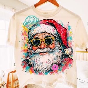 May include: Cream-coloured t-shirt featuring a colourful graphic of Santa Claus wearing sunglasses. The design includes the text "THE ORIGINAL" in a circular stamp. The Santa illustration has a white beard and a red hat, with colourful paint splatters.