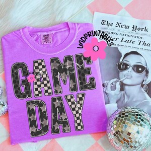 May include: A lilac-colored t-shirt with the words "GAME DAY" in a distressed font. The letters feature a mix of star and checkered patterns. A vintage-style image of a woman and a disco ball are in the background.
