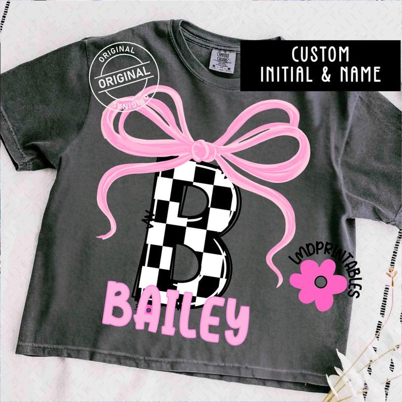 May include: Dark gray t-shirt with a pink bow and a large letter "B" in a black and white checkered pattern. The name "Bailey" is printed in pink below the letter. The shirt also features the text "CUSTOM INITIAL & NAME".