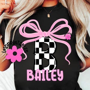 May include: Black t-shirt featuring a large pink bow and a checkered "B" in the center. The name "Bailey" is printed in pink below the letter. A pink flower graphic is on the left side.