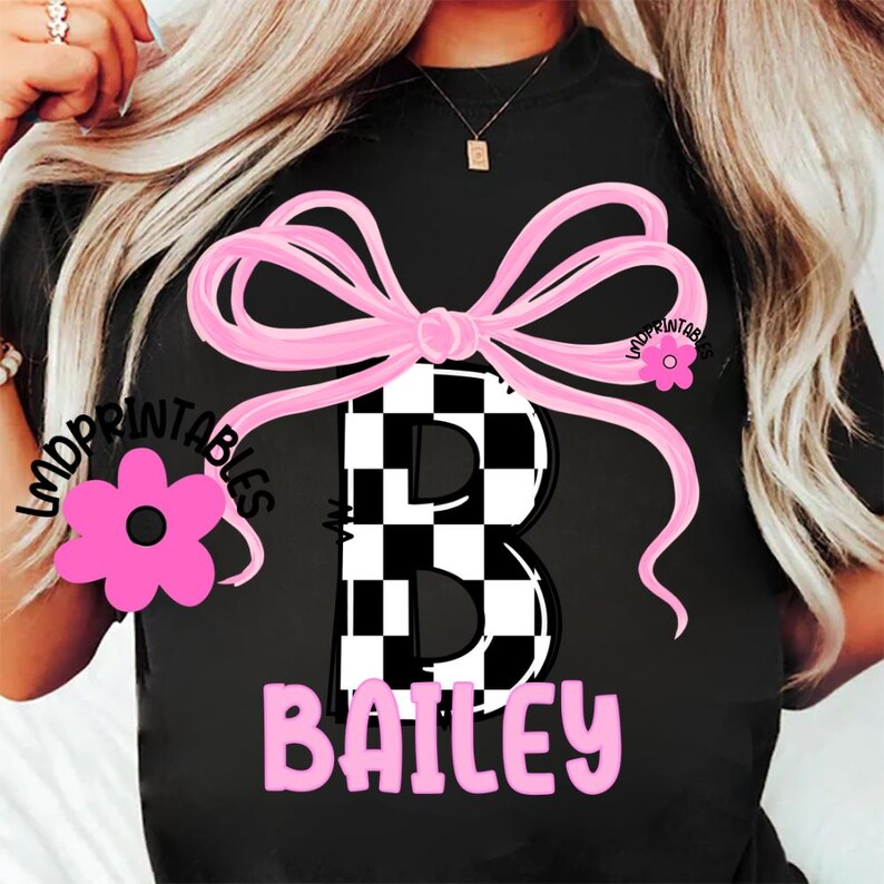 May include: Black t-shirt featuring a large pink bow and a checkered "B" in the center. The name "Bailey" is printed in pink below the letter. A pink flower graphic is on the left side.
