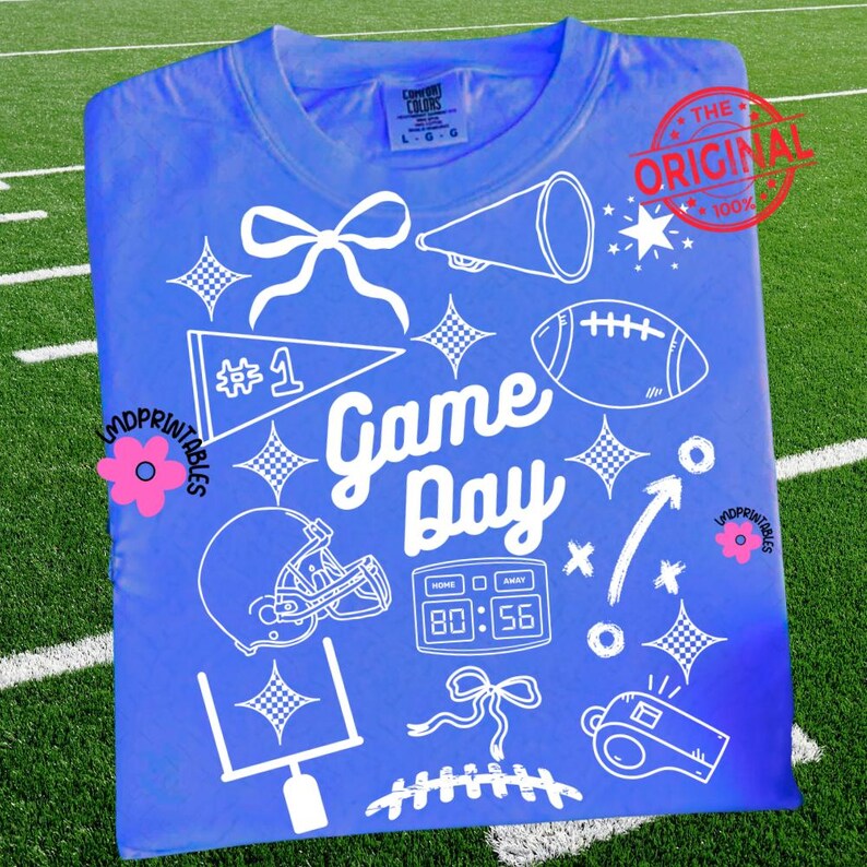 May include: A blue t-shirt with a white graphic design featuring football-related elements. The design includes a football, helmet, megaphone, and the words "Game Day." The shirt also has a "#1" pennant and a scoreboard graphic.
