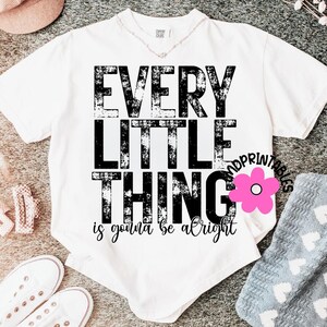 May include: White t-shirt with the text "EVERY LITTLE THING is gonna be alright" in black distressed font. A pink flower graphic is on the right side of the shirt. The shirt is on a gray surface with other clothing items.