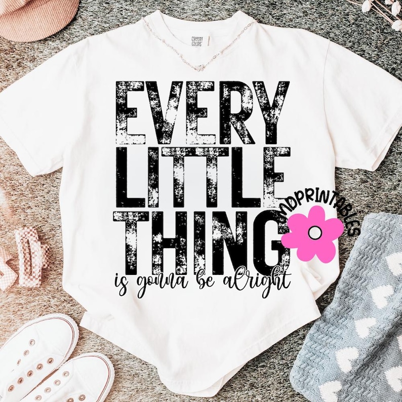 May include: White t-shirt with the text "EVERY LITTLE THING is gonna be alright" in black distressed font. A pink flower graphic is on the right side of the shirt. The shirt is on a gray surface with other clothing items.