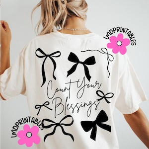 K&ouml;nnte beinhalten: Cremefarbenes T-Shirt mit schwarzen Schleifendesigns und dem handgeschriebenen Schriftzug "Count Your Blessings". Das Design umfasst verschiedene Schleifenstile und wird durch pinkfarbene Blumengrafiken mit dem Text "LMDPRINTABLES" akzentuiert.