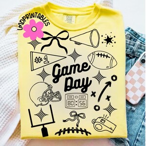 May include: Yellow t-shirt with black graphic elements including a football, helmet, and the words "Game Day." The design also features a pennant, a megaphone, and a scoreboard displaying 80:56. The shirt is a Comfort Colors brand.