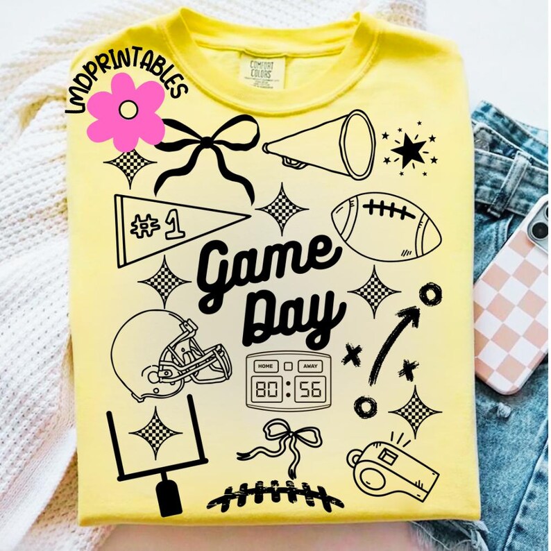May include: Yellow t-shirt with black graphic elements including a football, helmet, and the words "Game Day." The design also features a pennant, a megaphone, and a scoreboard displaying 80:56. The shirt is a Comfort Colors brand.