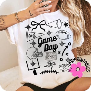 May include: White t-shirt with black graphic elements related to American football. The design includes a football, helmet, megaphone, and the words "Game Day". Additional elements include a bow, stars, and a scoreboard.