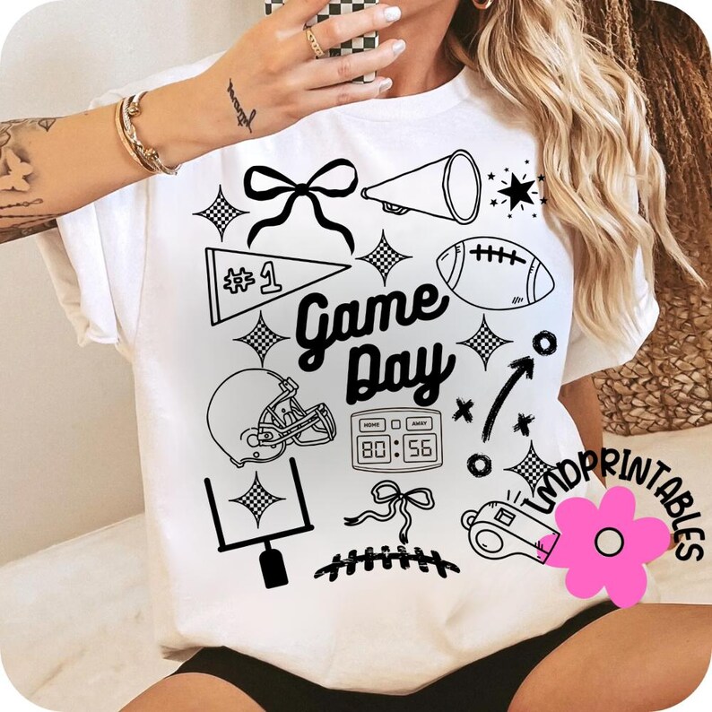 May include: White t-shirt with black graphic elements related to American football. The design includes a football, helmet, megaphone, and the words "Game Day". Additional elements include a bow, stars, and a scoreboard.