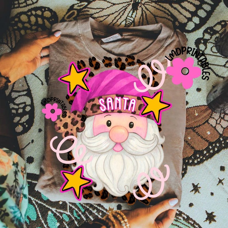 Op de afbeelding: Een beige T-shirt met een Santa Claus-afbeelding met een roze hoed en witte baard. Het woord "SANTA" staat op de hoed. Het ontwerp bevat gele sterren en roze bloemaccenten, met luipaardprintdetails.