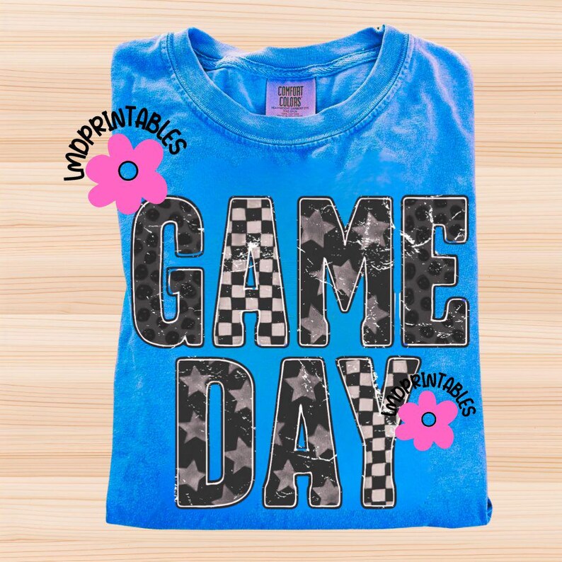 May include: A blue t-shirt with the words "GAME DAY" in a distressed, textured font. The letters feature checkered, star, and leopard print patterns. The shirt has a Comfort Colors tag and a pink flower graphic.