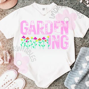 May include: White t-shirt with the text "GARDENING" in pink, featuring colorful flower graphics. The shirt has a crew neck and short sleeves. A necklace is visible at the neckline. The shirt is laying on a gray surface.