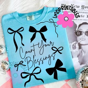 K&ouml;nnte beinhalten: Hellblaues T-Shirt mit schwarzen Schleifenmotiven und dem Schriftzug "Count Your Blessings" in Schreibschrift. Das Shirt hat einen Rundhalsausschnitt und kurze &Auml;rmel. Eine rosa Blumengrafik und ein Schwarz-Wei&szlig;-Bild einer Frau im Hintergrund.