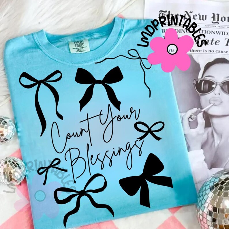 K&ouml;nnte beinhalten: Hellblaues T-Shirt mit schwarzen Schleifenmotiven und dem Schriftzug "Count Your Blessings" in Schreibschrift. Das Shirt hat einen Rundhalsausschnitt und kurze &Auml;rmel. Eine rosa Blumengrafik und ein Schwarz-Wei&szlig;-Bild einer Frau im Hintergrund.