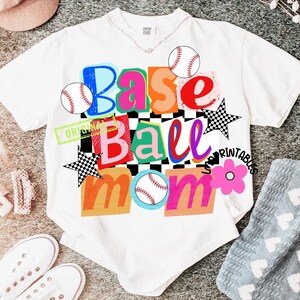 以下が含まれることがあります： 「Base Ball Mom」と書かれたカラフルなグラフィックが特徴の白いTシャツ。デザインには野球ボール、星、花が含まれています。テキストはさまざまな色とフォントで、チェック柄の背景です。
