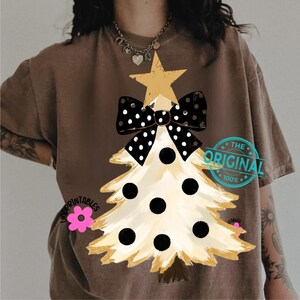 May include: Brown t-shirt featuring a Christmas tree design. The tree is cream-colored with black polka dots and a gold star and bow. The text "THE ORIGINAL 100%" is printed on the shirt.