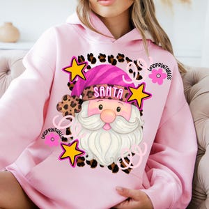 Op de afbeelding: Roze hoodie met een Santa Claus-afbeelding. Het Santa-ontwerp bevat een roze muts, witte baard en het woord "SANTA". Het ontwerp is omgeven door gele sterren, luipaardprint accenten en de tekst "LMDPRINTABLES".