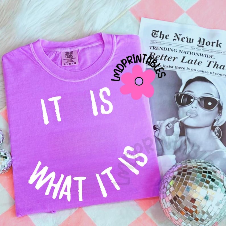 May include: A lilac-colored t-shirt with the phrase "IT IS WHAT IT IS" in white block letters. The shirt is displayed with a vintage newspaper and a disco ball. The shirt is a Comfort Colors brand.