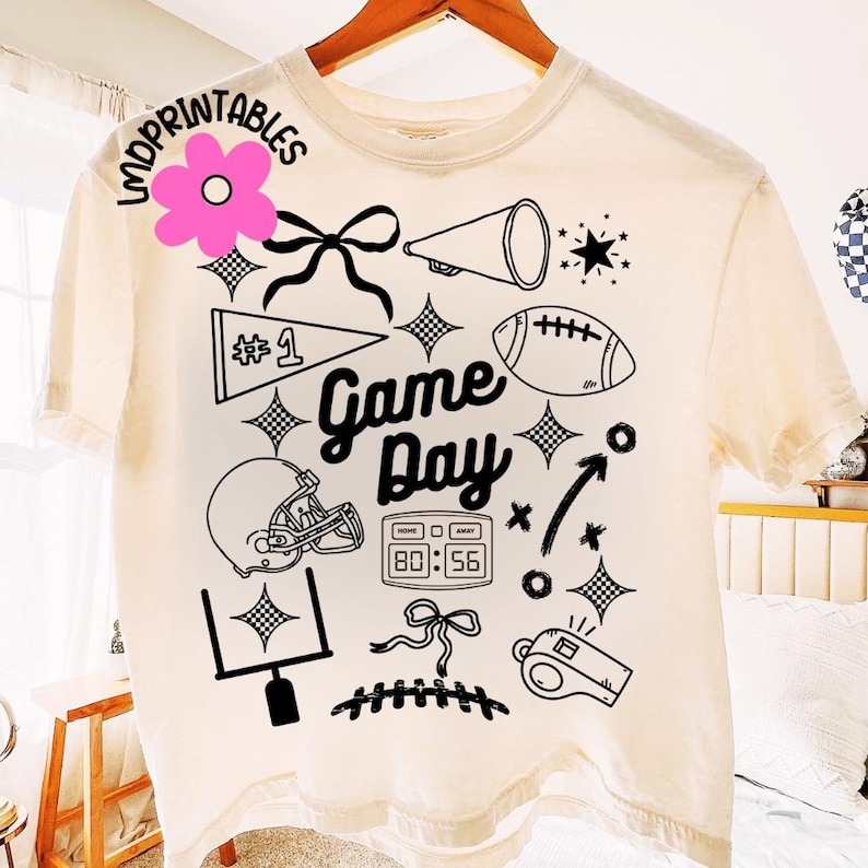 May include: Cream-colored t-shirt with a black graphic design featuring football-themed elements. The design includes a football, helmet, megaphone, and the words "Game Day". The shirt also has a score display and a whistle.