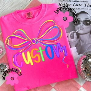 May include: Bright pink t-shirt with a colorful bow design and the word "CUSTOM" in rainbow letters. The shirt is a Comfort Colors brand. The background includes a newspaper clipping and disco ball.