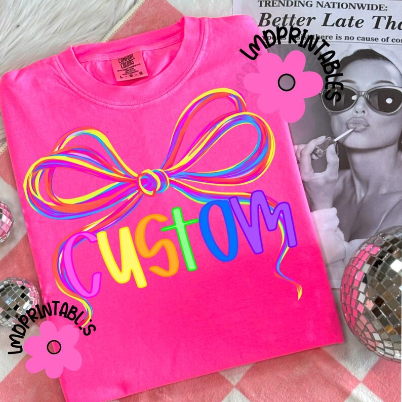 May include: Bright pink t-shirt with a colorful bow design and the word "CUSTOM" in rainbow letters. The shirt is a Comfort Colors brand. The background includes a newspaper clipping and disco ball.