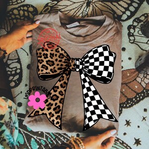 May include: A tan-colored t-shirt featuring a large bow design. One side of the bow has a leopard print, while the other is a black and white checkerboard pattern. A pink flower is below the bow, and the text "LADYPRINTABLES" is visible.