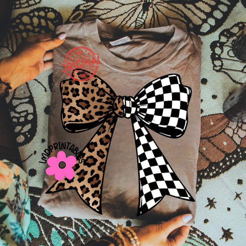 May include: A tan-colored t-shirt featuring a large bow design. One side of the bow has a leopard print, while the other is a black and white checkerboard pattern. A pink flower is below the bow, and the text "LADYPRINTABLES" is visible.